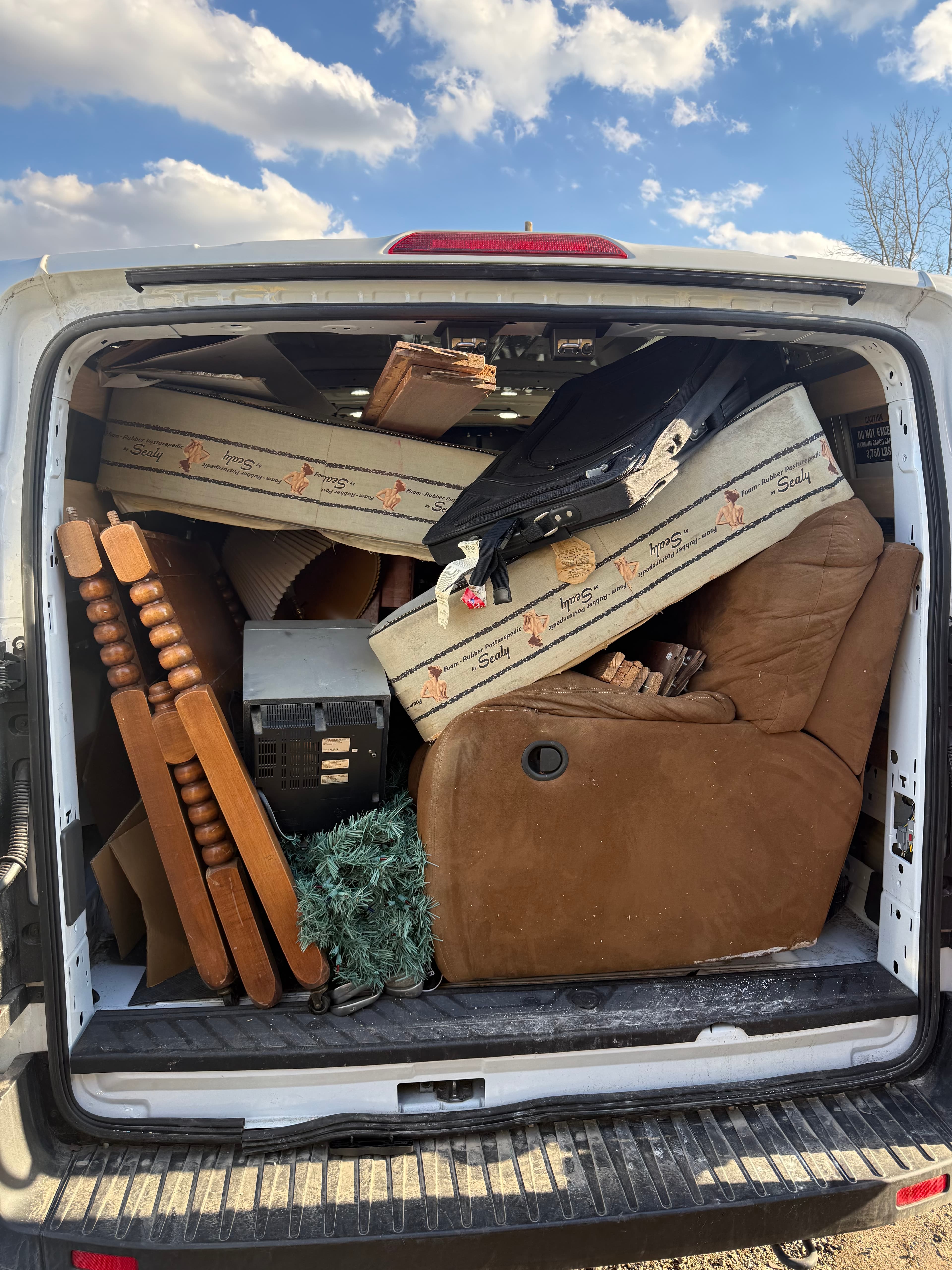 White van's open back packed with a brown recliner, Sealy mattresses, and wooden furniture.