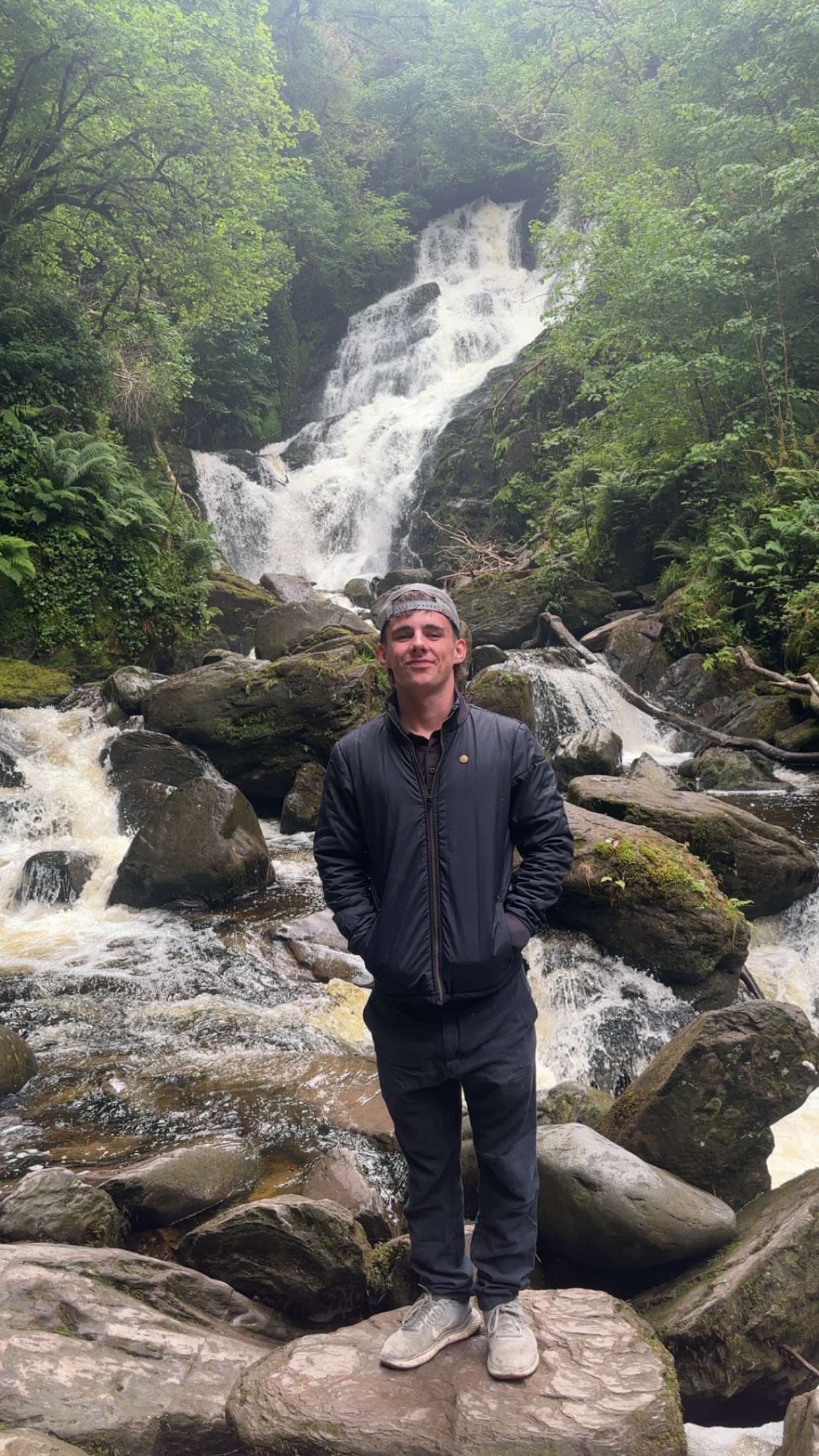 Smiling man in a dark jacket stands on rocks before a large cascading forest waterfall.